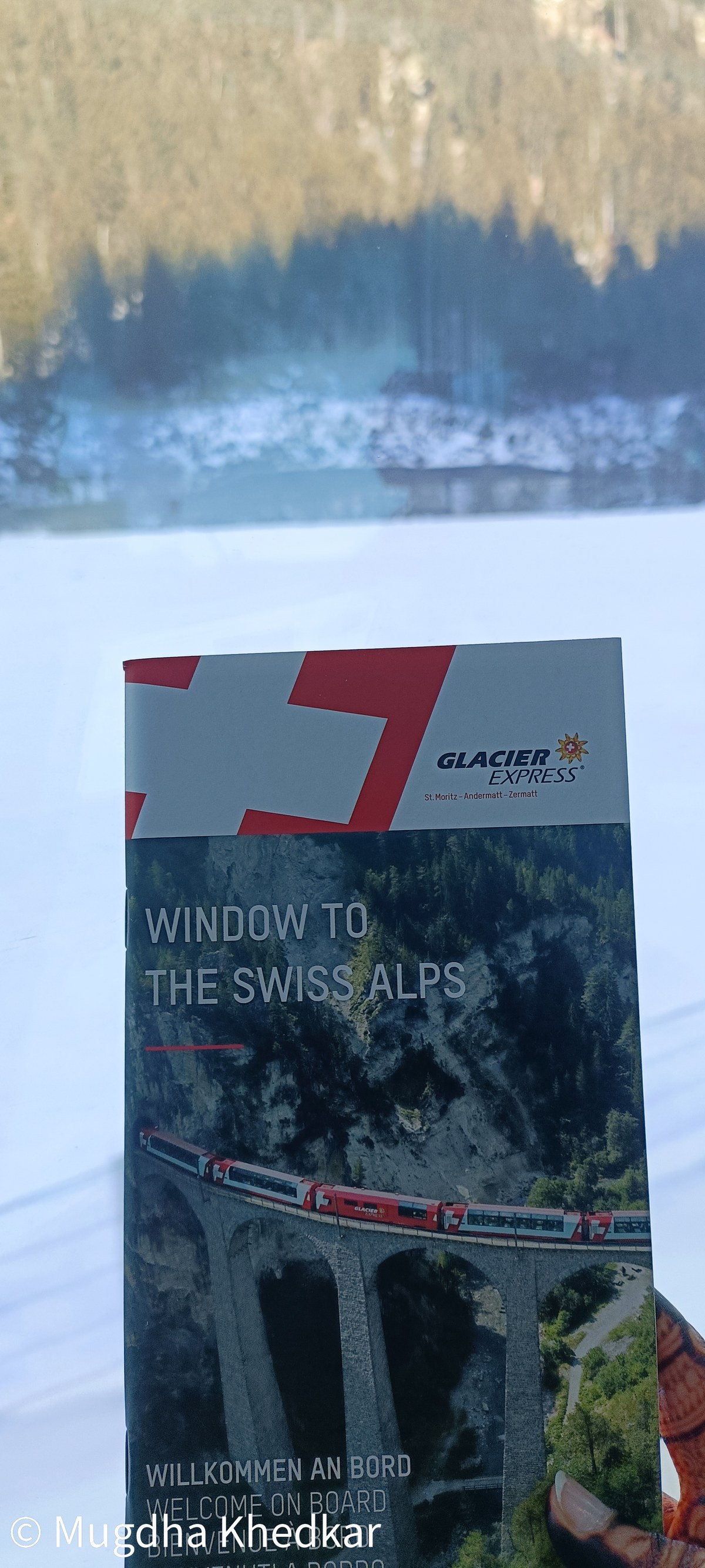 A Glacier Express brochure held against the train window with the snowy Alps outside